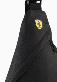 Sac bandoulière noir à surface texturée, présentant un motif losange et un logo bouclier jaune proéminent avec un cheval noir. Bretelle réglable.