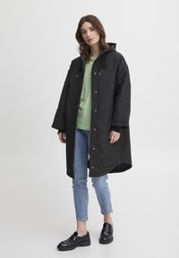 Sort kviltet lang frakke med hætte, trykknapper og justerbare snore; parret med en lys grøn skjorte, blå jeans og sorte sko.