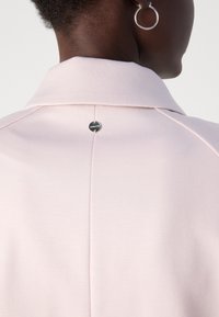 Lichtroze jasje met een gestructureerde kraag, voorzien van een centrale naad en een ronde zilveren logoaccent aan de achterkant. Gladde stoftextuur.