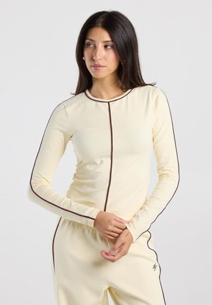 Vrouw met lang donker haar, gekleed in een crèmekleurige top met lange mouwen en broek met donkere biezen, kijkt opzij met gevouwen handen.
