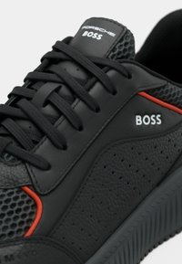 Baskets en cuir noir avec une surface texturée, des accents en mesh, des détails rouges et des lacets noirs. Présente le logo "BOSS" sur le côté.