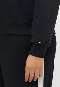 Sweatshirt noir avec des poignets côtelés, arborant un petit logo rouge, blanc et bleu sur la manche. Le tissu semble doux et texturé.