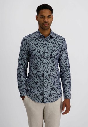 Mann trägt blaues, langärmeliges Hemd mit dunklem Blumenmuster und helle beige Hose, steht vor hellem einfarbigem Hintergrund.