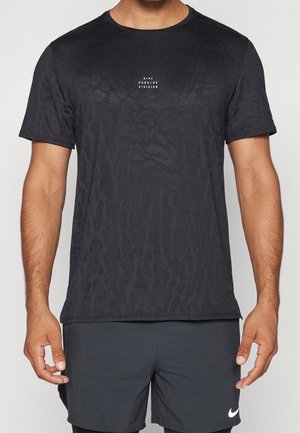 Mann trägt schwarzes Nike Running Division T-Shirt mit dezentem Muster und schwarze Shorts mit weißem Nike-Logo, steht mit entspannten Armen.
