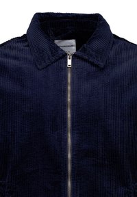 Veste en velours côtelé bleu marine avec une fermeture éclair argentée à l'avant et un col plié, montrant la partie supérieure de la poitrine et du cou.
