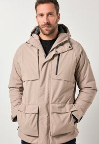 Uomo che indossa un giubbotto invernale beige con cappuccio e molte tasche a pattina, con un maglione nero sotto, mani in tasca.