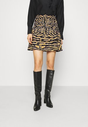 Vrouw draagt een zwarte top met lange mouwen, een minirok met tijgerprint en zwarte kniehoge laarzen met hakken, staand tegen een effen achtergrond.
