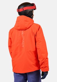 Helderef oranjerode skijack met een capuchon, met een gladde textuur en logodetail op de achterkant, gedragen over blauwe skibroek en handschoenen.