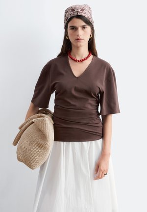 Kvinde iført brun V-hals skjorte, hvid nederdel, rød perlekæde, mønstret hovedtørklæde, der holder stor strikket beige taske mod ensfarvet baggrund.