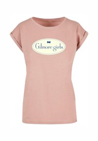 Rosa kortermet T-skjorte med rullet ermer. Har en hvit oval logo med blå tekst "Gilmore girls" og en blå sommerfugl som accent.