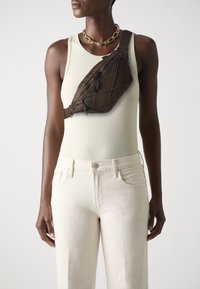 Brown, triangular crossbody bag with a textured finish, featuring a zippered compartment and adjustable strap, worn over a light tank top.