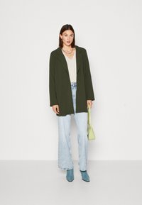 Olive green blazer over a light V-neck top, paired with light blue wide-leg jeans and blue ankle boots. A small green handbag accents the outfit.