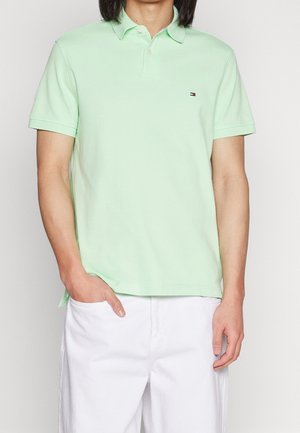 Hombre con polo de manga corta verde claro con un pequeño logo en el pecho y pantalones blancos, con una mano en el bolsillo.