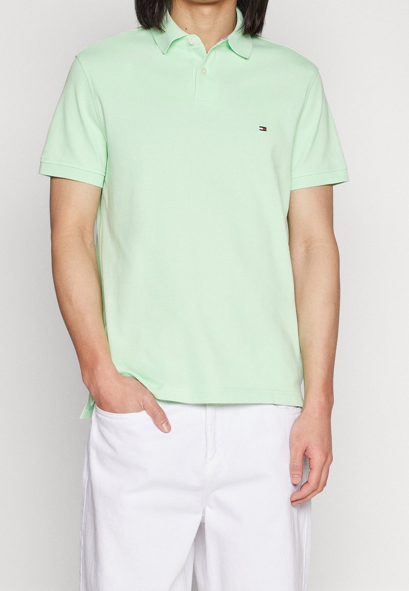 Homme portant un polo à manches courtes vert clair avec un petit logo sur la poitrine et un pantalon blanc, une main dans la poche.