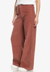 Pantalons en velours côtelé à jambes larges couleur rouille, avec des côtes verticales, une taille haute et une fermeture à bouton à l'avant. Des baskets blanches complètent le look.