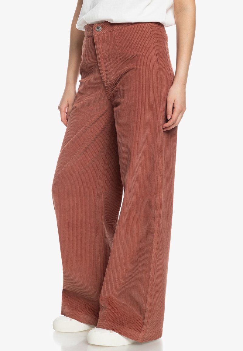 Pantalons en velours côtelé à jambes larges couleur rouille, avec des côtes verticales, une taille haute et une fermeture à bouton à l'avant. Des baskets blanches complètent le look.