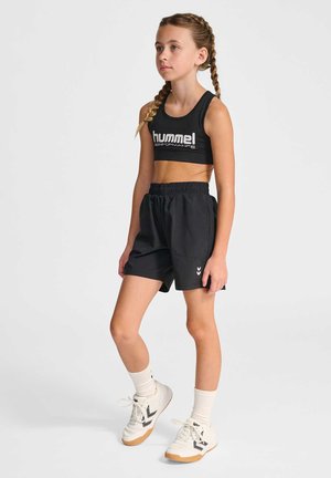Junges Mädchen mit geflochtenem Haar, das einen schwarzen Sport-BH und Shorts, weiße Socken und weiße Sneaker trägt, steht vor einem schlichten Hintergrund.