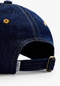 Căciulă din denim de culoare albastră închisă, cu cusături galbene, având o curea ajustabilă și un etichetă albă cu inscripția "SCALPERS" pe spate.