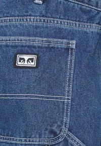 Dark blue denim fabric with white stitching, featuring a back pocket and a rectangular logo patch with a black border and white design.