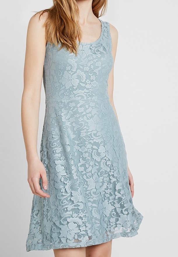 Femme portant une robe sans manches, longueur genoux, en dentelle bleu clair avec des motifs floraux sur un fond uni.