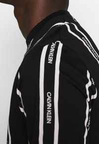 Černé tričko s krátkým rukávem a bílými svislými pruhy, na kterém je nápis "Calvin Klein" a žebrovaný výstřih.