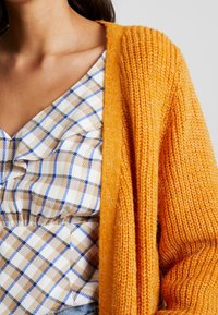 Frau trägt ein beiges, blaues und weißes karierte Oberteil mit Rüschen und einen orangefarbenen grob gestrickten Cardigan über einer Schulter.
