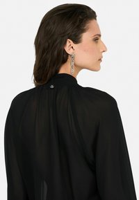 Blouse noire transparente avec un col haut et un doux plissé. Présente une fermeture à boutons à l'arrière et une élégante boucle d'oreille en chaîne métallique.