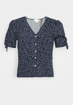 Blouse bleu marine à manches courtes avec des taches abstraites blanches, col en V, boutons sur le devant et poignets de manches noués.