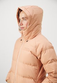 Podšívaná kabátik v broskyňovej farbe s vysokým golierom a pripevnenou kapucňou. Hladká látka s viditeľným vzorom šitia a zipsami.