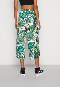 Spodnie o długości 7/8 z żywym zielonym i białym tropikalnym wzorem liści, wykonane z lekkiego materiału, w zestawieniu z czarnymi trampkami.