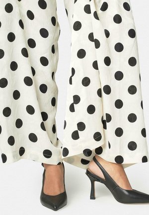 Mujer con pantalones blancos de pierna ancha con grandes lunares negros y zapatos slingback negros de punta puntiaguda con tacones de aguja.