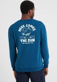 Man wearing blue long-sleeve shirt with white graphic of bird, sun, waves, and text "Here Comes The Sun Tog Twenty Four" on the back.
