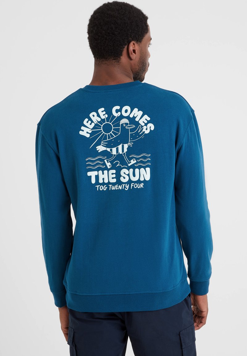 Man wearing blue long-sleeve shirt with white graphic of bird, sun, waves, and text "Here Comes The Sun Tog Twenty Four" on the back.