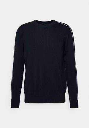 Mies seisoo käsineen taskuissa, päällään musta huppari ja siihen sopivat housut valkoisilla raitoilla, jalassaan valkoiset lenkkarit yksinkertaisella taustalla.