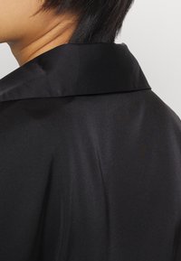 Černá saténová košile s širokým límcem, hladkým povrchem a jemným leskem, z pohledu zezadu, zvýrazňující elegantní design.