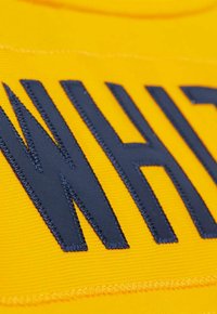 Nahaufnahme von schwarzen gestickten Buchstaben "WAS" auf einem strukturierten gelben Stoffhintergrund.