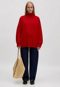 Roter, lässiger Strickpullover mit Rollkragen und geripptem Saum, kombiniert mit dunkelblauen weiten Jeans. Trägt eine beige, weiche Tote-Tasche. Schwarze Schuhe.