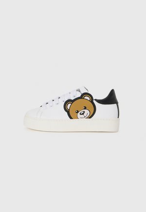 Vit sneaker med svarta accenter, med ett björnfacedesign på sidan. Slät material, rund tå och platt gummisula.