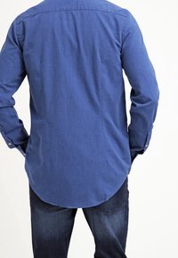 Camicia a maniche lunghe blu scuro con orlo curvo, polsini con bottone e dettaglio sulla parte posteriore, abbinata a jeans in denim scuro. Tessuto liscio.