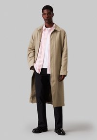 Abrigo largo beige con un corte relajado sobre una camisa de rayas rojas y blancas, superpuesta a una camiseta blanca. Pantalones y zapatos negros completan el look.