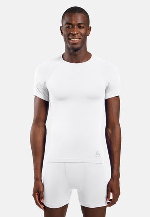 Homme souriant portant un t-shirt de sport blanc ajusté et un short, debout devant un fond clair uni.
