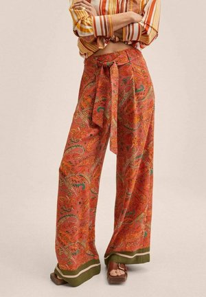 Frau trägt hoch taillierte, weit geschnittene Hose mit orangefarbenem Paisley-Muster und gebundener Taille, kombiniert mit braunen Plateausandalen und einem gestreiften Hemd.