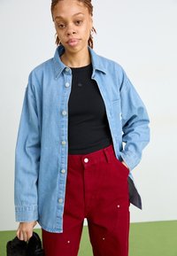 Denim košulja u svijetloplavoj boji s srebrnim dugmadi, složena preko crne rebraste košulje, uparena s tamnocrvenim hlačama koje imaju džepove i detalje od hardvera.