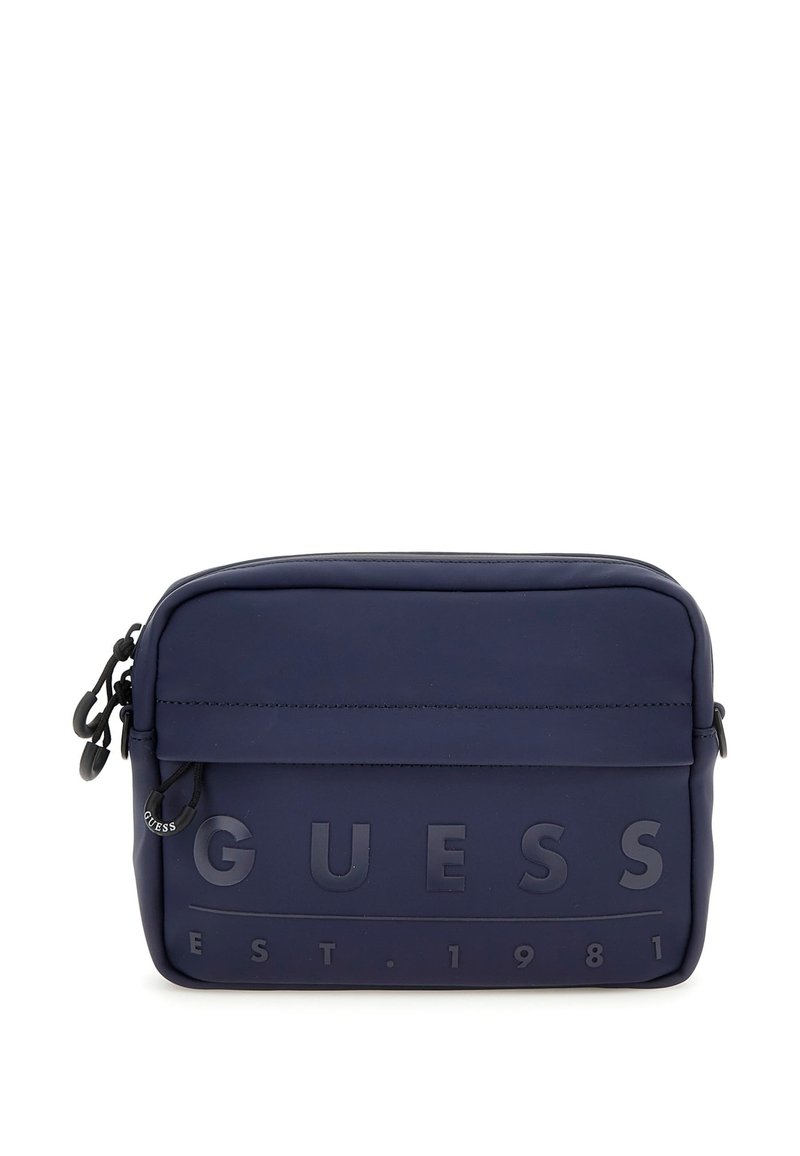 Bolsa de material suave en azul marino con el texto en relieve "GUESS EST. 1981", cierre con cremallera y correa ajustable. Textura suave con un diseño minimalista.