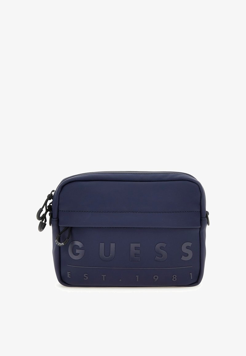 Bolsa de material suave en azul marino con el texto en relieve "GUESS EST. 1981", cierre con cremallera y correa ajustable. Textura suave con un diseño minimalista.