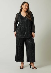 Zwart paillettentop en wijde pijpen broekset, met een V-hals, lange mouwen en een gestructureerde stof met een glanzende afwerking. Strappy zwarte hakken.
