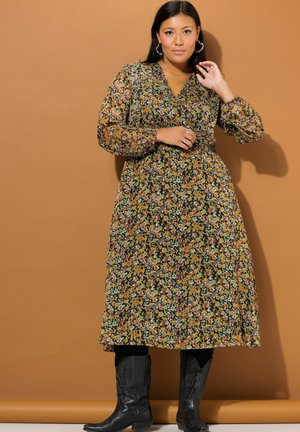 Una mujer posa frente a un fondo beige, vestida con un vestido largo de flores, medias negras, botas hasta la rodilla, pendientes de aro y un collar.