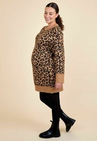 Tan ja musta леопардiprindiga kampsunimeel dress, rööbastega ääre ja varrukatega, koos mustade pahkluusaabaste ja mustade retuusidega. Lihtne taust.