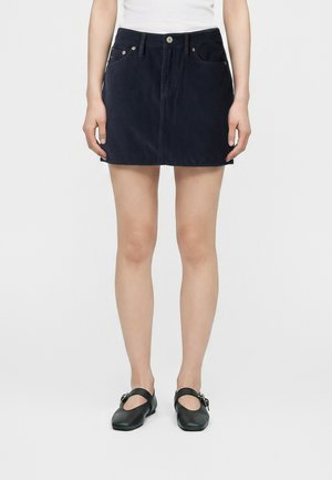 Femme portant une mini-jupe en velours côtelé bleu marine, un haut blanc et des chaussures noires style Mary Jane, debout devant un fond clair uni.