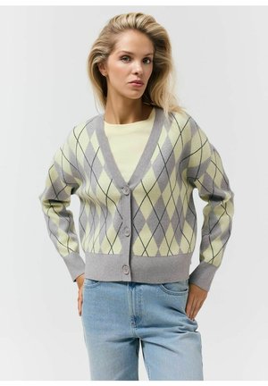 Blondynka w jasnym żółtym i szarym rozpinanym swetrze w romby na kremowej koszuli i jasnoniebieskich dżinsach, pozująca z rękami na biodrach.